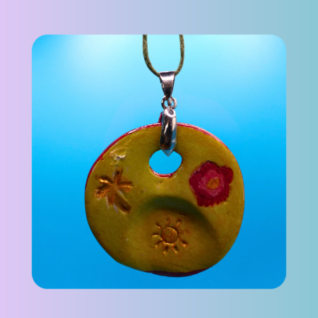 Vue de face du pendentif Petit Lien en argile autodurcissante, pièce unique, fait main, couleur jaune beige, forme organique ronde, surface douce et brillante. Posé sur fond bleu. Motifs fleur, palmier et soleil