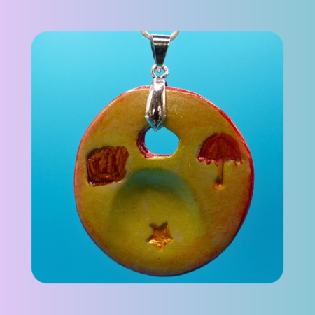 Vue de face du pendentif Petit Lien en argile autodurcissante, pièce unique, fait main, couleur sable, motifs parapluie, étoile et valise, forme organique ronde, surface douce et brillante. Posé sur fond bleu.