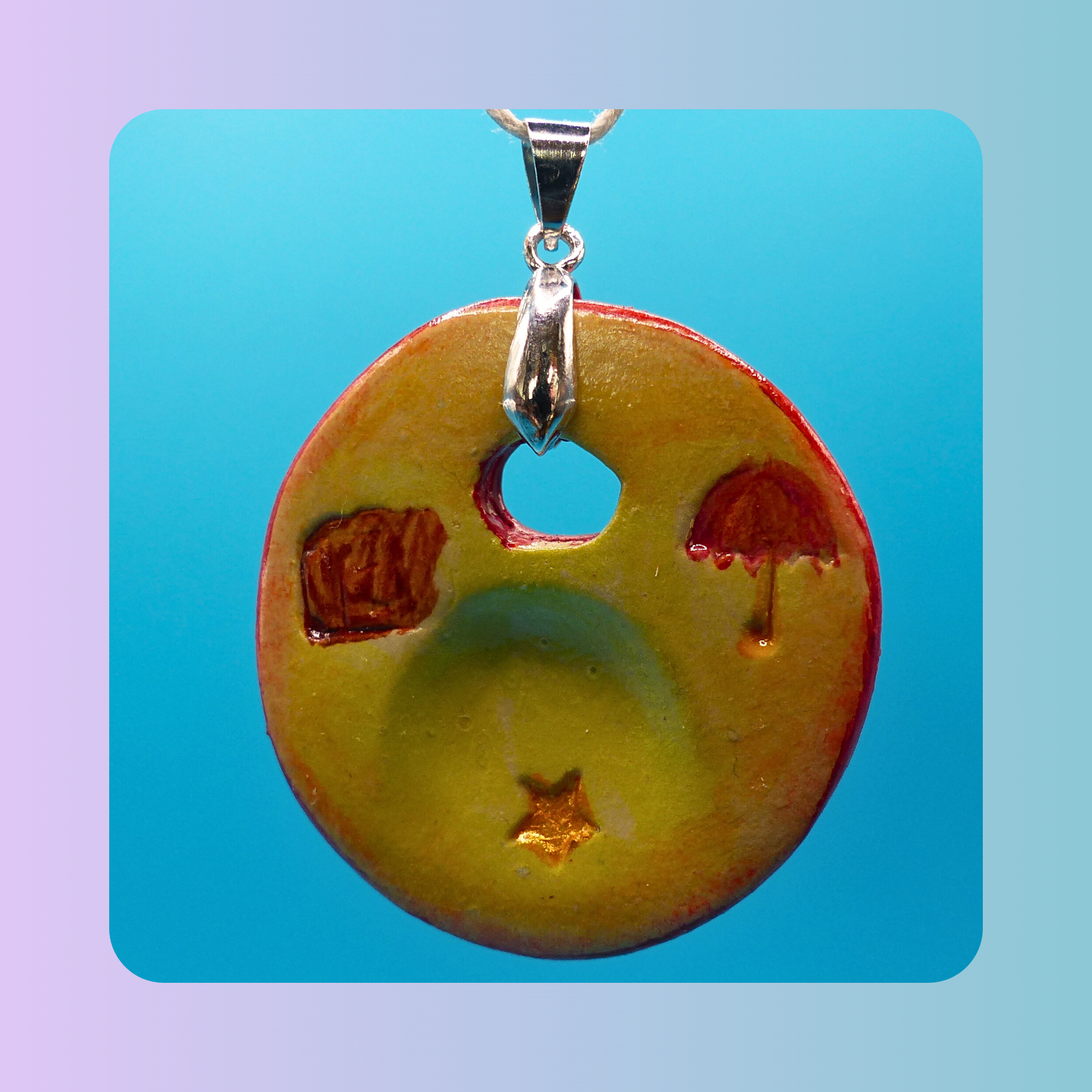 Vue de face du pendentif Petit Lien en argile autodurcissante, pièce unique, fait main, couleur sable, motifs parapluie, étoile et valise, forme organique ronde, surface douce et brillante. Posé sur fond bleu.