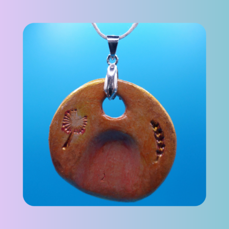 Vue de face du pendentif Petit Lien en argile autodurcissante, pièce unique, fait main, couleur orangée, forme organique ronde, surface douce et brillante. Motif de pissenlit et épi de blé. Posé sur fond bleu.