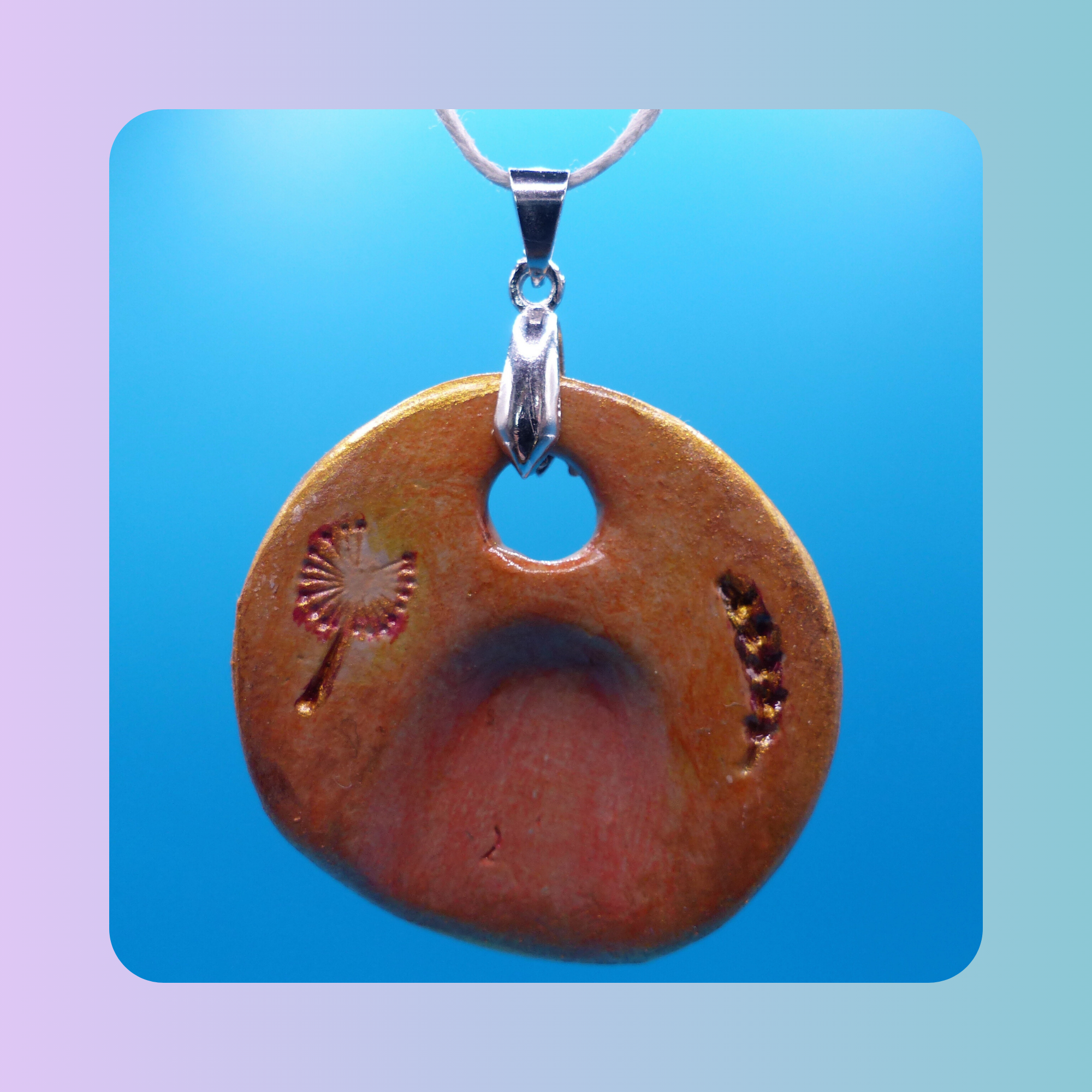 Vue de face du pendentif Petit Lien en argile autodurcissante, pièce unique, fait main, couleur orangée, forme organique ronde, surface douce et brillante. Motif de pissenlit et épi de blé. Posé sur fond bleu.