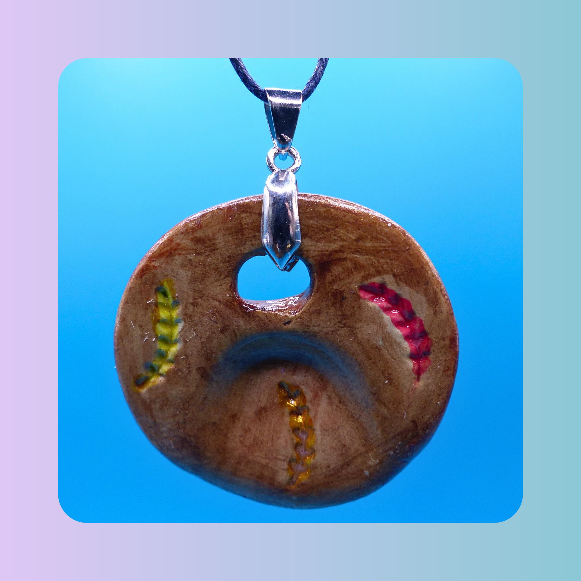 Vue de face du pendentif Petit Lien en argile autodurcissante, pièce unique, fait main, couleur, forme organique allongée ou ronde, surface douce et brillante. Posé sur fond bleu. Motif de trois épis de blé